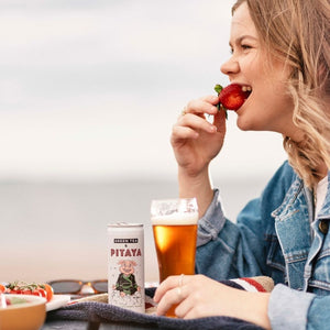 Blonde girl eating strawberry and smiling while sitting at table. Holding a glass of nitro cold brew green tea and pitaya. A can of East Forged with Notsu pig on the front smiling. There are some bowls of food and sunglasses nearby on the table.