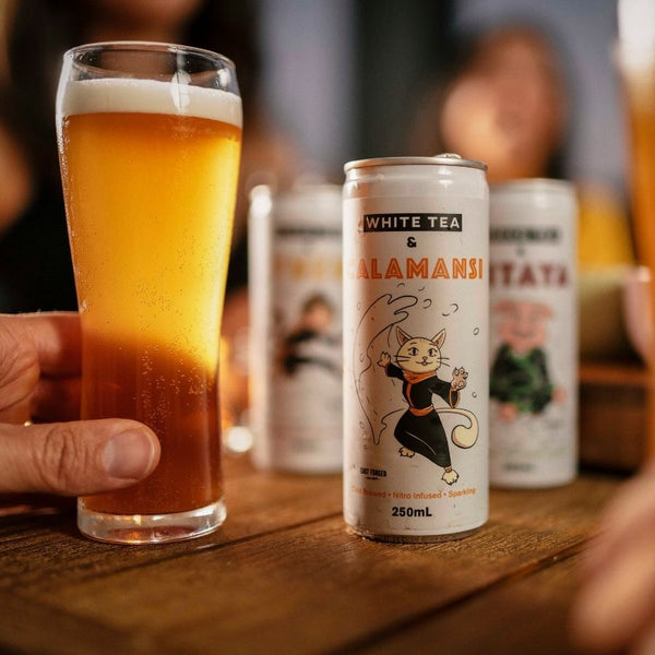 Hand holding a glass of nitro cold brew tea near the can with Saka cat on the front. In the background, blurry images of people and two other flavours of East Forged cans