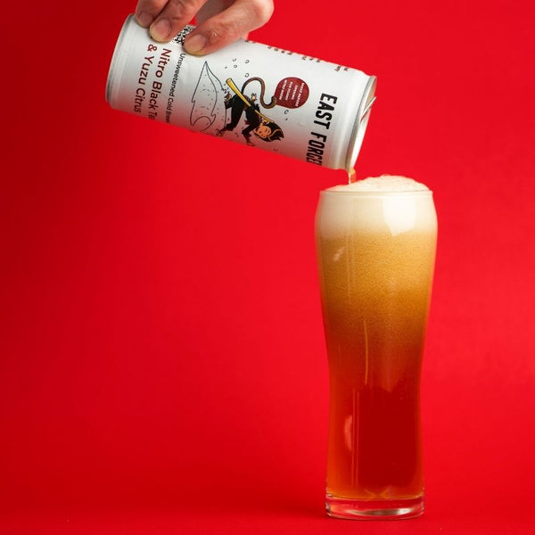 Bright red background, hand holding a can of East Forged nitro black tea & yuzu citrus with Mico Monkey on the front.  Pouring into glass almost full and nitro foam.