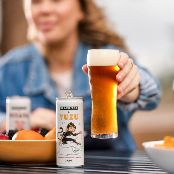 Smiling girl, blurry in the back, holding a clear image of a glass with nitro cold brew tea. She is sitting at a table with a bowl of oranges and berries. A can of East Forged black tea and yuzu with Mico smiling monkey is beside the glass