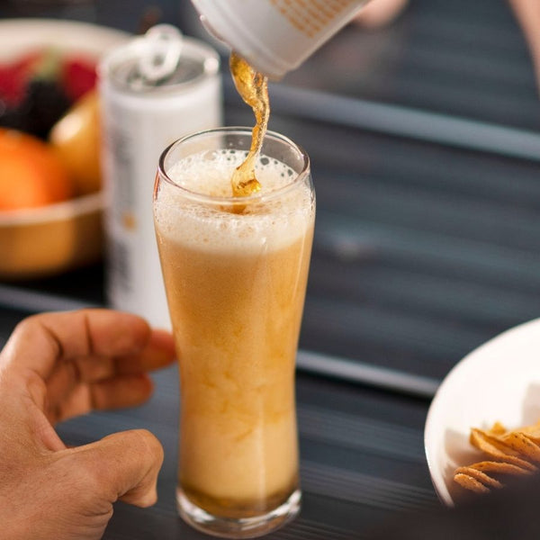 Pouring a can of black tea & yuzu into a glass.  The nitro foam is surging in the glass.  It is almost full.  There is a hand beside the glass and chips in a bowl.  There is a can and bowl of fruit blurry in the background.