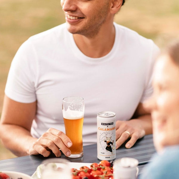 Smiling man sitting at table, holding glass of nitro cold brew white tea.  Near an East Forged can with Saka cat on the front.
