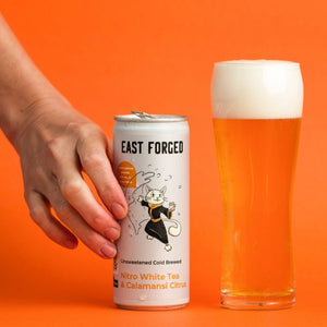 Hand holding a can of East Forged nitro white tea & calamansi citrus with Saka cat on the front.  Glass of poured tea and nitro foam beside the can. Bright orange background.